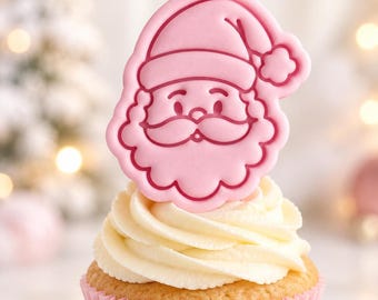 May include: A cupcake in a light pink paper liner topped with white swirled frosting and a flat, light pink Santa Claus face decoration. The Santa topper features a detailed embossed outline of a hat, beard, mustache, and eyes on a smooth surface.