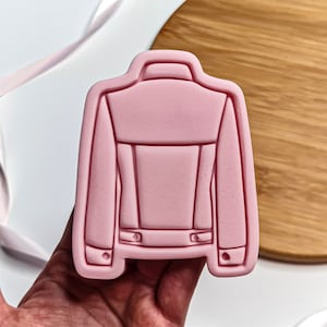 May include: Pink plastic cookie cutter shaped like the back of a denim jacket.