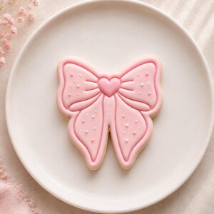 May include: A bow-shaped sugar biscuit decorated with light pink icing, featuring a central heart detail and small white dot accents. The biscuit is centred on a plain white plate, showcasing smooth icing lines and a defined, symmetrical shape.