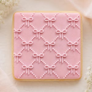 May include: Square-shaped cookie with a pink icing design featuring a pattern of bows and dotted lines. The cookie has a light brown edge and is set against a soft, light background with white flowers.