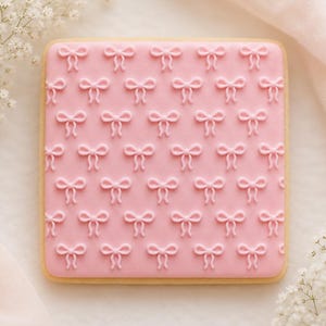 May include: A square-shaped cookie with a light brown edge and a pink icing top. The pink icing is decorated with a repeating pattern of small, raised bow shapes. The cookie is presented on a white surface with flowers.