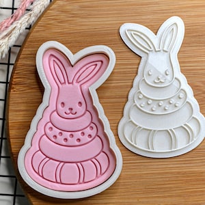 Puede incluir: Un cortador de galletas de plástico blanco en forma de conejo que lleva una pila de anillos. El cortador se muestra junto a una galleta rosa que se ha cortado con el cortador.