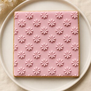 May include: A square sugar cookie with a smooth, light pink icing finish. The surface is decorated with a repeating, embossed pattern of small, eight-petaled flowers in a slightly darker shade of pink, arranged in a uniform grid across the cookie.