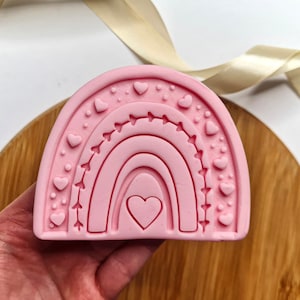 May include: Pink silicone mold shaped like a rainbow with a heart in the center. The rainbow has a dotted and heart-shaped border.