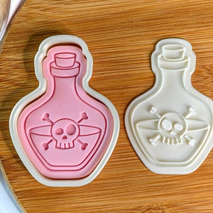 Puede incluir: Dos cortadores de galletas de plástico blanco con forma de botellas de poción con un diseño de calavera y huesos cruzados. El cortador de la izquierda está lleno de fondant rosa.