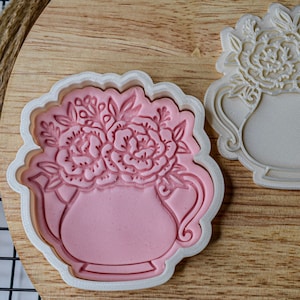 Puede incluir: Dos cortadores de galletas de plástico blanco con forma de tetera con flores en la parte superior. El cortador de la izquierda tiene fondant rosa en su interior.