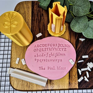 May include: A set of alphabet stamps for baking and crafting. The stamps are white and come in a yellow container. The stamps are arranged on a pink fondant with the alphabet printed in black. The text "The Pink Ideas" is printed on the fondant.