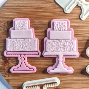 Puede incluir: Dos cortadores de galletas de pastel de fondant rosa con un diseño de encaje. Los cortadores están sobre una superficie de madera.