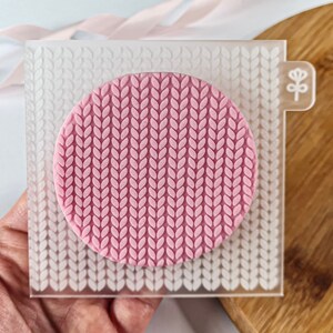 May include: A clear silicone stamp with a raised design of a knitted pattern. The stamp is pink and has a small flower design in the corner.