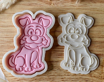 May include: A white plastic cookie cutter in the shape of a cartoon dog holding a bone in its mouth. The cutter is shown with a pink impression of the dog.