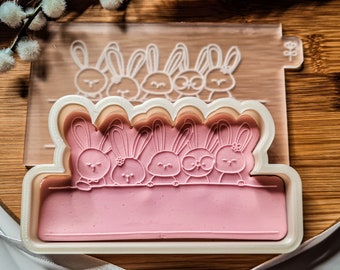 May include: A white plastic cookie cutter with a pink background. The cutter is shaped like a banner with five bunnies peeking over the top. The bunnies have black eyes and noses and are outlined in black.