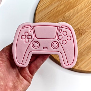 May include: Pink plastic cookie cutter shaped like a video game controller. The controller has buttons and a directional pad.