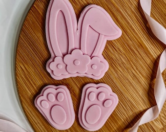 Op de afbeelding: Roze fondant konijnenoren en poot afdrukken koekjesvormen. De konijnenoren hebben drie bloemen aan de onderkant.