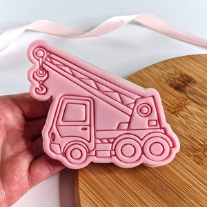 May include: Pink plastic cookie cutter shaped like a crane truck. The cutter has a detailed design with a hook, cab, and wheels.