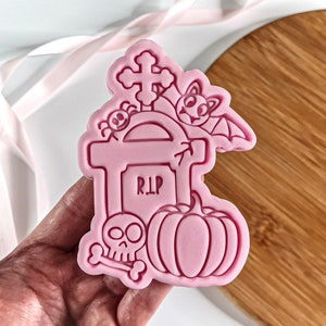 May include: Pink silicone Halloween cookie cutter featuring a tombstone, cross, bat, pumpkin, and skull and crossbones design. The tombstone is inscribed with "RIP".