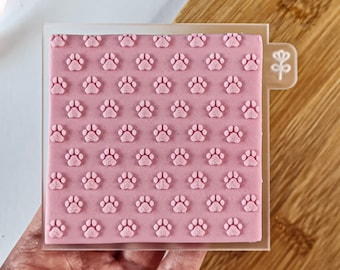 May include: A clear acrylic stamp with a pink paw print design. The stamp is on a wooden surface.
