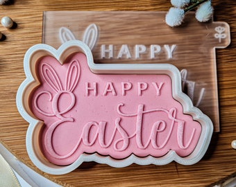 Op de afbeelding: Een witte plastic koekjesvormer met een roze ontwerp met de tekst "Happy Easter" met konijnenoren.