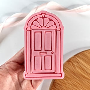 May include: Pink plastic cookie cutter shaped like a traditional front door with a fanlight window above the door. The door has a doorknob, a letterbox, and two panels.