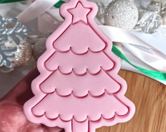 May include: A pink Christmas tree cookie cutter with a star detail. The cutter features a detailed design with scalloped edges and a rectangular base. The background includes white and silver festive decorations.