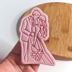 Puede incluir: Cortador de galletas de plástico rosa en forma de novia y novio besándose. La novia lleva un ramo de flores.
