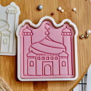 May include: Two white plastic cookie cutters shaped like a mosque. One cutter is filled with pink modeling clay, showing the outline of the mosque.