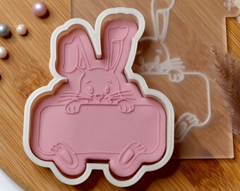May include: White plastic cookie cutter shaped like a bunny rabbit holding a blank sign. The bunny has pink ears and a pink nose.
