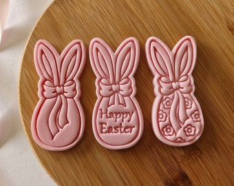May include: Three pink bunny-shaped cookie cutters, one with the text "Happy Easter" on the front, one with a bow and a floral design, and one with a bow.