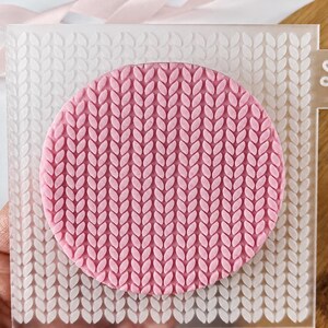 May include: A square, translucent acrylic embossing plate featuring a repeating knit stitch pattern. A circular pink textured disc with the same knit pattern is centered on the plate. A small flower icon is etched on the right tab of the plate.