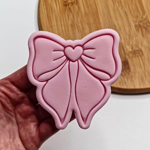 Puede incluir: Cortador de galletas de plástico rosa en forma de lazo con un corazón en el centro.