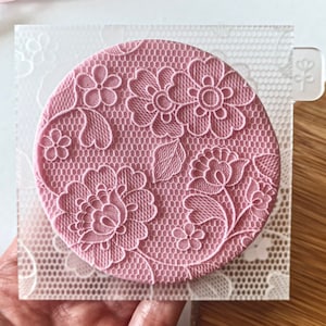 Könnte beinhalten: Eine transparente Plastikfolie mit einer rosa Silikonform eines runden Spitzenmusters. Die Form zeigt ein zartes Blumenmuster mit Herzen und Blättern.