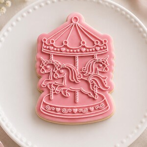 May include: A sugar cookie shaped like a carousel with a pink icing base and white piped details. The design features three unicorns on poles under a canopy, with small heart accents along the roof and the base of the ride, presented on a white plate.