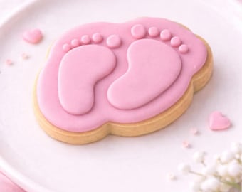 May include: A pink baby footprint biscuit on a white plate. The biscuit has a light brown base with pink icing showing two baby footprints. Small pink heart-shaped sweets and flowers surround the biscuit.
