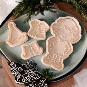 Puede incluir: Un conjunto de cortadores de galletas de plástico beige con forma de elfo, sombrero de elfo, camisa de elfo y botas de elfo, dispuestos sobre un plato azul claro. También hay una galleta de chocolate decorada y ramitas de pino.