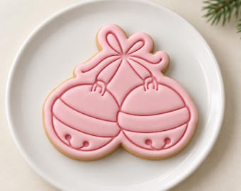 May include: A light pink sugar cookie shaped like two jingle bells tied together with a bow. The cookie features embossed details of the bells and the bow, with a smooth icing finish covering the entire surface of the cookie shape.