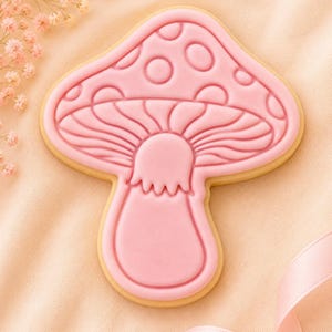 Puede incluir: Una galleta con forma de seta decorada con glaseado rosa claro liso. El glaseado presenta detalles en relieve, incluyendo un sombrero redondeado con manchas circulares y un tallo con un anillo ondulado. La galleta tiene un borde beige.