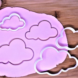 May include: Four white plastic cloud-shaped cookie cutters on a pink surface. The cutters are of different sizes and are arranged in a row.