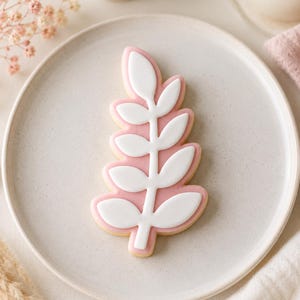May include: A leaf-shaped sugar biscuit decorated with smooth pink icing as the base and a white icing overlay in the shape of a central stem with five pairs of symmetrical, rounded leaves extending outwards, centred on a round ceramic plate.