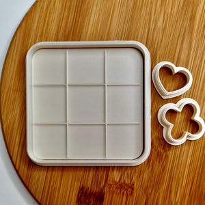 May include: A white plastic cookie cutter set with a square grid design, a heart shape, and a four-leaf clover shape.