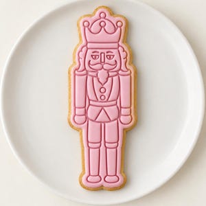 May include: A nutcracker-shaped sugar cookie decorated with smooth, light pink icing. The icing features embossed details of a crown, a mustache, a uniform with buttons, and tall boots, all outlined in a slightly darker shade of pink icing.