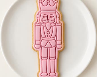May include: A nutcracker-shaped sugar cookie decorated with smooth, light pink icing. The icing features embossed details of a crown, a mustache, a uniform with buttons, and tall boots, all outlined in a slightly darker shade of pink icing.