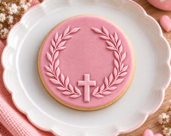 May include: A round sugar cookie topped with smooth pink icing, featuring a raised relief design of a laurel wreath encircling a central cross. The cookie is centered on a white scalloped plate, showcasing a clean, symmetrical decorative pattern.