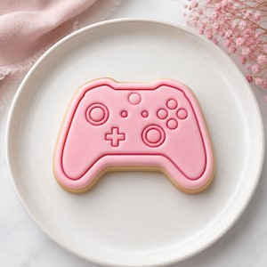Puede incluir: Una galleta con forma de mando de videojuego, con una capa de glaseado rosa claro liso y detalles en relieve que incluyen dos joysticks, una cruceta direccional y cuatro botones circulares, sobre un plato redondo de color blanco.