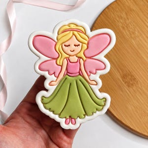 May include: A decorated sugar cookie shaped like a fairy with blonde hair, a pink top, and a green skirt. The fairy has pink wings and closed eyes, outlined in white icing. The design features smooth, colorful icing details on a white base.