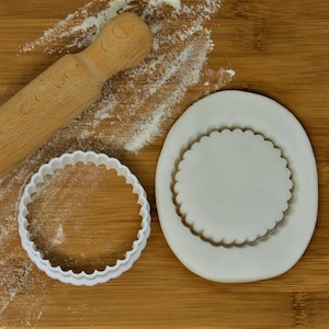 May include: Two white plastic cookie cutters on a wooden surface. One cutter is round with a scalloped edge and the other is a plain circle. There is a wooden rolling pin and white flour on the surface.