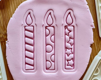 May include: Three pink birthday candle cookie cutters with different designs: stripes, polka dots, and stars.