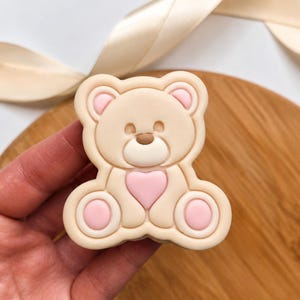 May include: A beige teddy bear shaped cookie cutter with a raised design featuring a small pink heart on the chest, pink circular accents on the feet, and a brown nose. The cutter has a thick, moulded plastic frame with a rounded, smooth edge.