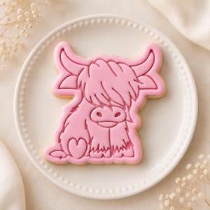May include: A light pink biscuit shaped like a Highland cow with shaggy hair, curved horns, and a small heart detail on its side. The biscuit features embossed lines defining the facial features and fur texture, centred on a white plate.