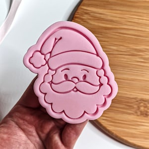 Könnte beinhalten: Ein rosa Ausstecher in Form eines Weihnachtsmann-Gesichts. Der Ausstecher hat ein detailliertes Design des Gesichts des Weihnachtsmanns, einschließlich Hut, Schnurrbart und Bart. Der Ausstecher wird in einer Hand gehalten, mit einem Holzschneidebrett im Hintergrund.