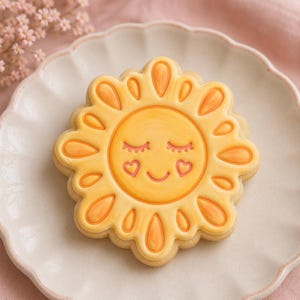 May include: A round sugar biscuit decorated with yellow and orange icing in the shape of a sun. The sun features a smiling face with closed eyes, pink eyelashes, and two small pink heart-shaped cheeks, surrounded by stylised sun rays.