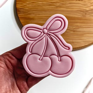 Può includere: Tagliapasta in plastica rosa a forma di due ciliegie con un fiocco in cima.